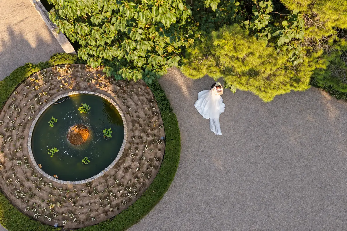 Novia junto a una fuente circular en un jardín vista desde un dron