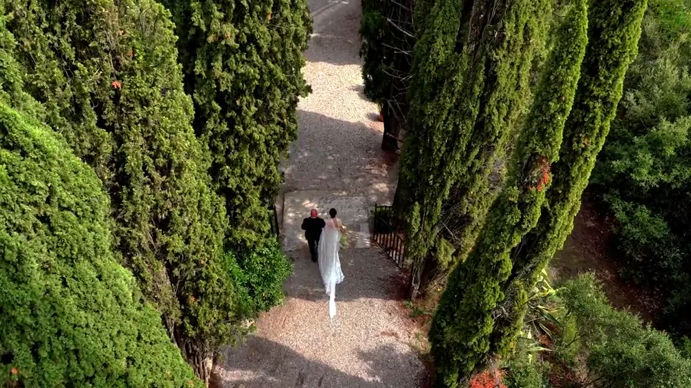 Novios caminando entre cipreses hacia una ceremonia en un palacete, vista desde un dron