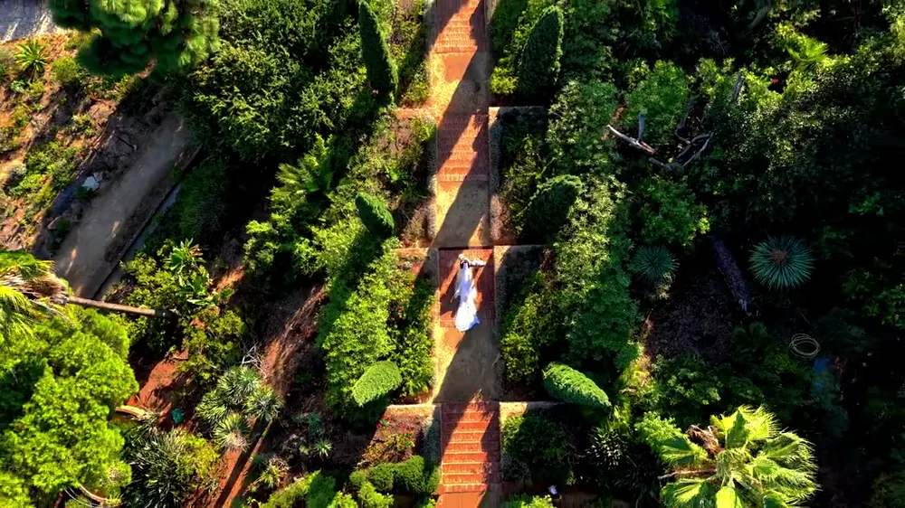 Novios en una escalera de jardín y una torre sobre el mar, vistos desde un dron