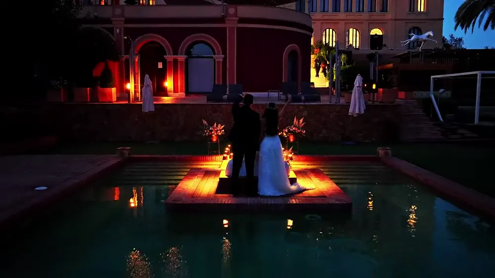 Novios frente a una piscina iluminada y un palacio al anochecer, vistos desde un dron