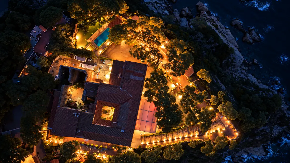 Hotel iluminado de noche sobre un acantilado de la Costa Brava, visto desde un dron